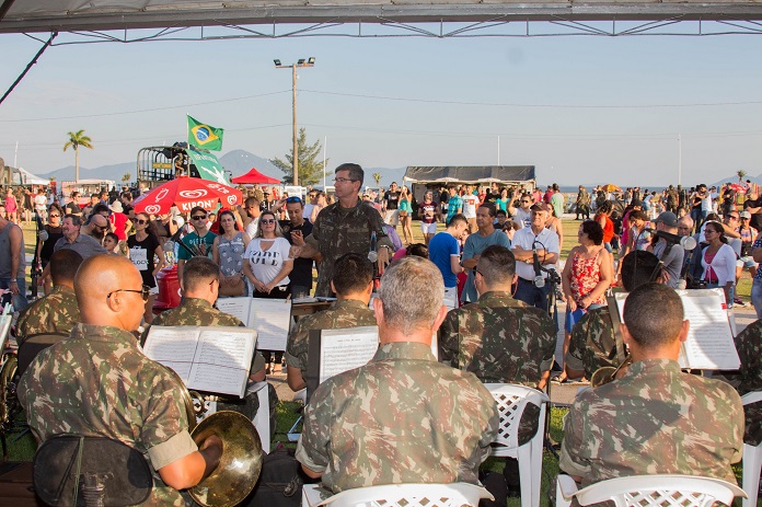 homens com roupa camuflada do exército tocando em uma orquestra; maestro em pé de frente para câmera rege a música e populares ao fundo observam