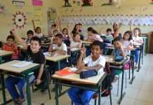 crianças sentadas em carteiras na sala de aula com os braços levantados
