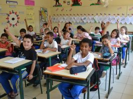 crianças sentadas em carteiras na sala de aula com os braços levantados