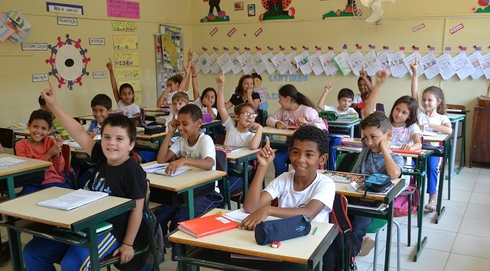 crianças sentadas em carteiras na sala de aula com os braços levantados