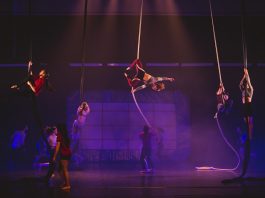 Florianópolis recebe 1ª etapa do Festival Internacional de Circo cenário de um espetáculo em um palco iluminado de roxo com artistas circulando; uma mulher está pendurada em uma grande fita que pende do teto