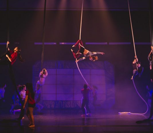 cenário de um espetáculo em um palco iluminado de roxo com artistas circulando; uma mulher está pendurada em uma grande fita que pende do teto