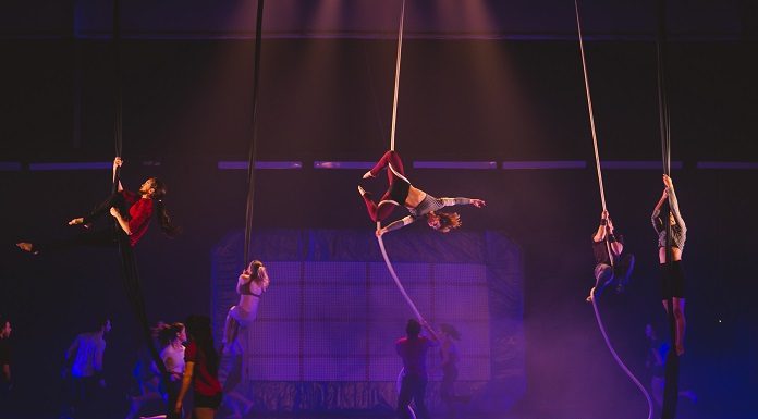 cenário de um espetáculo em um palco iluminado de roxo com artistas circulando; uma mulher está pendurada em uma grande fita que pende do teto