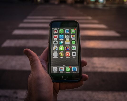 close em uma mão que segura um celular com tela ligada mostrando aplicativos; pessoa está em frente a uma faixa de pedestre