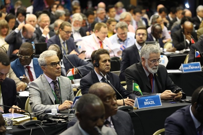 três homens membros da comissão sentados em bancada com a placa do brasil à frente ouvem fone de ouvido cercado por dezenas de outras pessoas, também membros de bancadas de outros países
