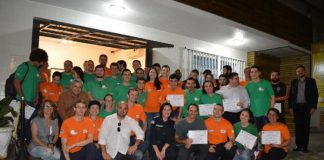 grupo de voluntários, com camisas laranjas ou verdes, posa para a foto de noite. ao centro do grupo e agachada está a secretária de segurança de florianópolis