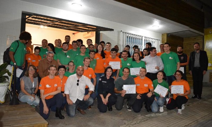 grupo de voluntários, com camisas laranjas ou verdes, posa para a foto de noite. ao centro do grupo e agachada está a secretária de segurança de florianópolis