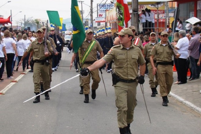 policiais militares em linha, carregando bandeiras, e um marchando na frente com uma espada, em uma avenida, observados por populares