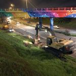 foto tirada de cima de um barranco mostra uma retroescavadeira abrindo buraco ao lado da rodovia; outros operários e máquinas circulam pelo acostamento, sob um viaduto colorido