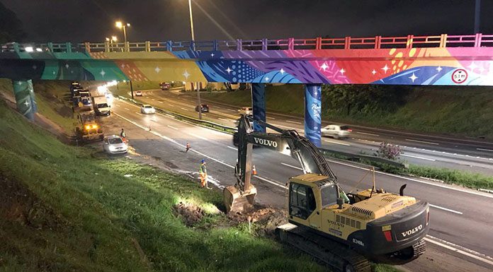 foto tirada de cima de um barranco mostra uma retroescavadeira abrindo buraco ao lado da rodovia; outros operários e máquinas circulam pelo acostamento, sob um viaduto colorido