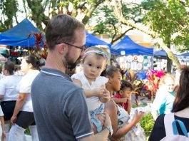 Feira da Freguesia festeja a Primavera em edição neste domingo (9) menina bebê no colo de um homem olha para a câmera; ao redor pessoas circulam e barracas da feira ao fundo, com algumas árvores