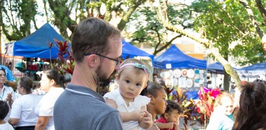Feira da Freguesia festeja a Primavera em edição neste domingo (9) menina bebê no colo de um homem olha para a câmera; ao redor pessoas circulam e barracas da feira ao fundo, com algumas árvores