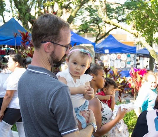 Feira da Freguesia festeja a Primavera em edição neste domingo (9) menina bebê no colo de um homem olha para a câmera; ao redor pessoas circulam e barracas da feira ao fundo, com algumas árvores