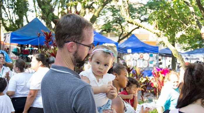 menina bebê no colo de um homem olha para a câmera; ao redor pessoas circulam e barracas da feira ao fundo, com algumas árvores