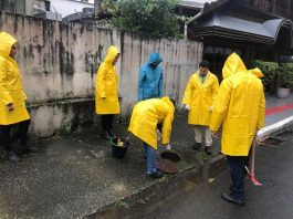 em um dia nublado, homens com capas de chuva amarelas estão em pé parados em volta de um bueiro olhando para o chão