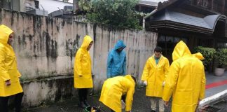 em um dia nublado, homens com capas de chuva amarelas estão em pé parados em volta de um bueiro olhando para o chão