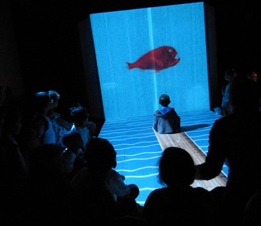 crianças sentadas no chão assistem a uma peça em ambiente muito escuro iluminado apenas ao centro onde há um peixe