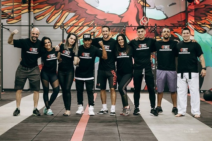 homens e mulheres da equipe da academia, com mesmo uniforme preto, posam em pé abraçados e sorridentes para a foto