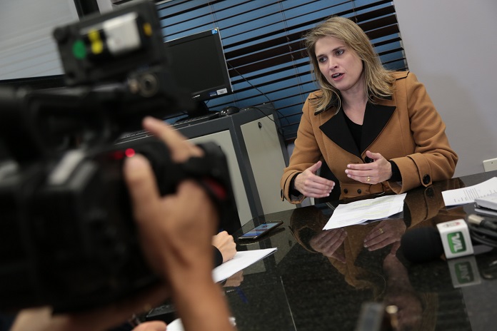 defensora dá entrevista sentada à frente de uma mesa; há um homem com uma câmera no quanto esquerdo da foto