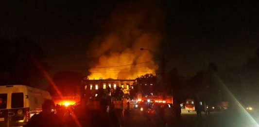 foto noturna da fachada do prédio histórico em chamas; algumas silhuetas de agentes de segurança na frente