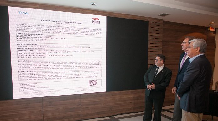 os três homens em pé ao lado de uma grande tela projetada na parede mostrando um documento de licença ambiental