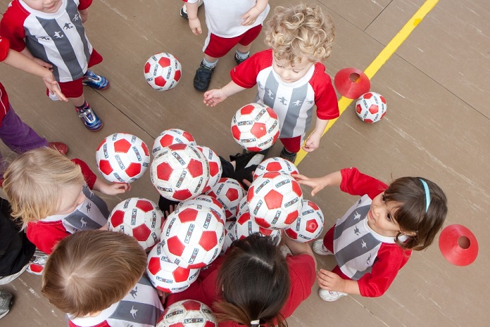 crianças pequenas com o mesmo uniforme tentam pegar bolas de um monte, vistas de cima em uma quadra
