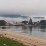 praia com mar calmo e enseada ao fundo com nevoeiro sobre o continente