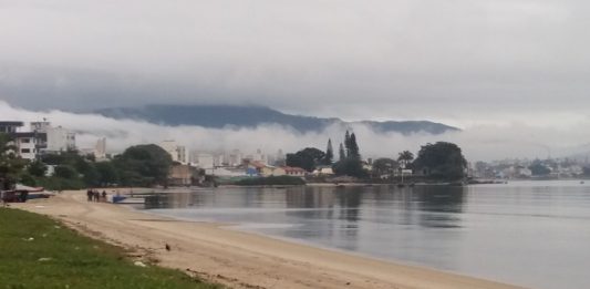 praia com mar calmo e enseada ao fundo com nevoeiro sobre o continente