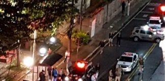 foto tirada do alto de um edifício mostra muitas viaturas paradas em volta de corpo no chão, com muitos policiais, ao lado de uma praça, onde populares olham