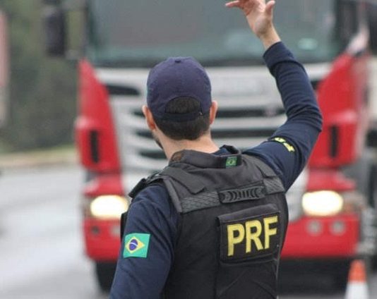 agente da prf visto de costas com uniforme da corporação gesticula com o braço direito para caminhão na estrada