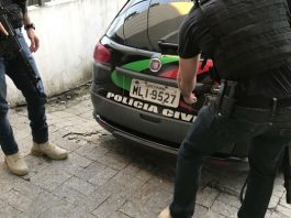 Polícia Civil localiza novas vítimas de quadrilha de estelionatários em Palhoça dois policias com metralhadoras em pé ao lado de carro da polícia civil; um deles se abaixa para alcançar a tampa do porta mala