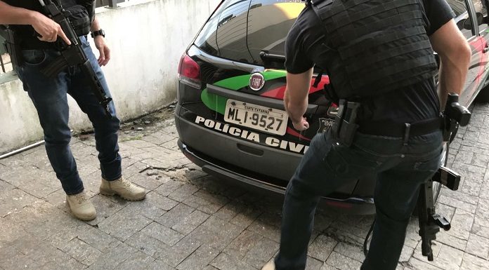 dois policias com metralhadoras em pé ao lado de carro da polícia civil; um deles se abaixa para alcançar a tampa do porta mala