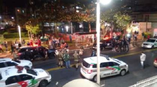 foto tirada do alto de um edifício mostra muitas viaturas paradas em volta de corpo no chão, com muitos policiais, ao lado de uma praça, onde populares olham