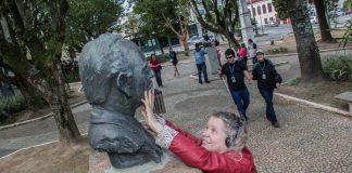 mulher passa as mãos no rosto de um busto em um praça; ao fundo, outros deficientes visuais caminham pelo espaço