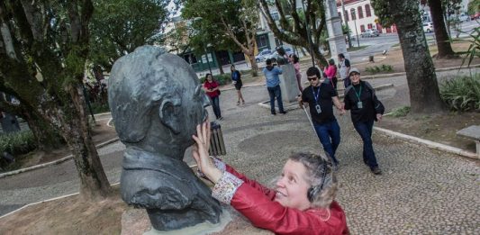 mulher passa as mãos no rosto de um busto em um praça; ao fundo, outros deficientes visuais caminham pelo espaço