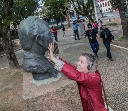 mulher passa as mãos no rosto de um busto em um praça; ao fundo, outros deficientes visuais caminham pelo espaço