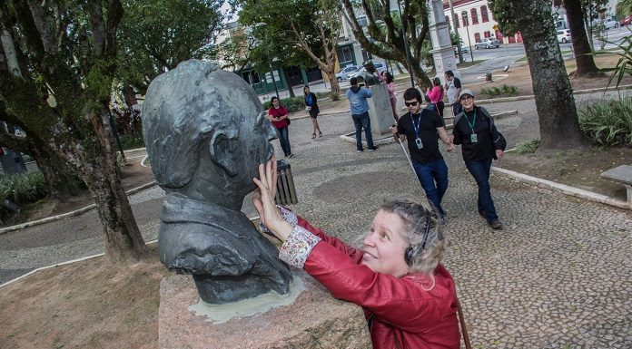 mulher passa as mãos no rosto de um busto em um praça; ao fundo, outros deficientes visuais caminham pelo espaço