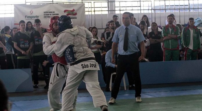 dois competidores de taekwondo lutam, um com capacete azul, outro vermelho, observados pelo juiz no tatame e público em geral na arquibancada em volta