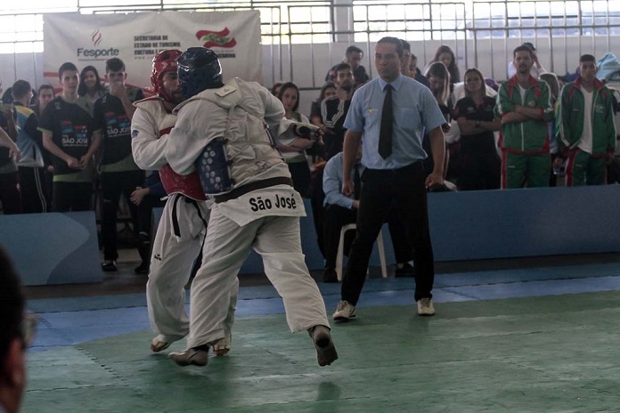 dois competidores de taekwondo lutam, um com capacete azul, outro vermelho, observados pelo juiz no tatame e público em geral na arquibancada em volta