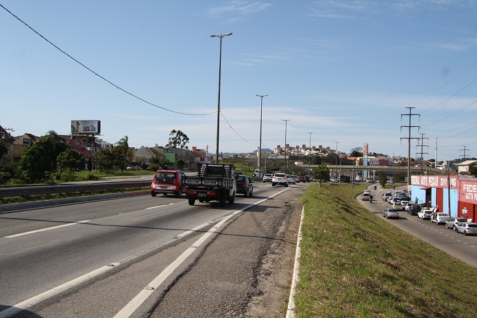 carros passam pela rodovia em foto tirada no acostamento em dia ensolarado