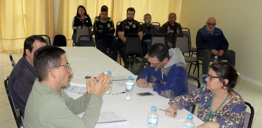 em uma mesa onde estão duas pessoas de cada lado, homem gesticula conversando com uma mulher à sua frente; ao fundo da sala, pessoas assistem sentadas em cadeiras lado a lado