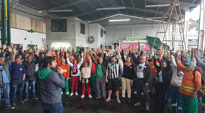 diversas pessoas com o uniforme da comcap com as mãos levantadas para cima em votação; há um homem à frente com um microfone; estão sob um galpão