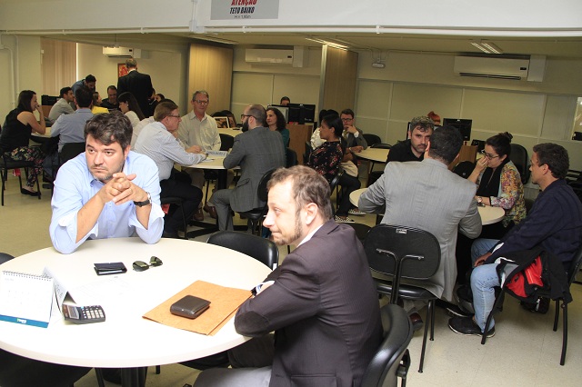 diversas pessoas sentadas em mesas redondas em uma grande sala; em primeiro plano, dois homens em uma mesa olham para o mesmo lado