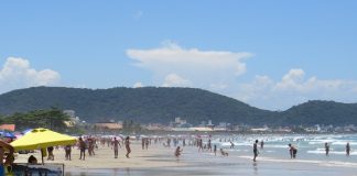 praia larga em um dia ensolarado; pessoas são vistas na distância circulando; algumas em primeiro plano debaixo de guarda sol