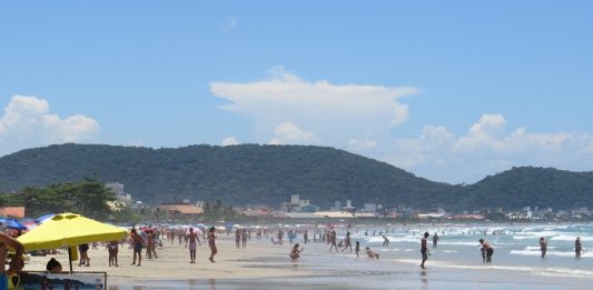 praia larga em um dia ensolarado; pessoas são vistas na distância circulando; algumas em primeiro plano debaixo de guarda sol