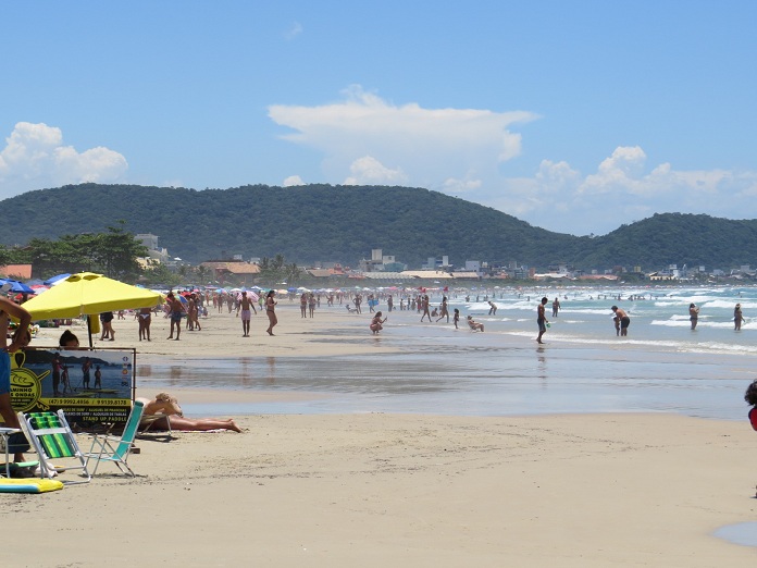 praia larga em um dia ensolarado; pessoas são vistas na distância circulando; algumas em primeiro plano debaixo de guarda sol
