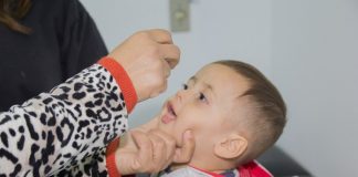 garotinho abre a boca para receber gotas de vacinação, segurado por uma mulher adulta; só se vê os braços e mãos dela