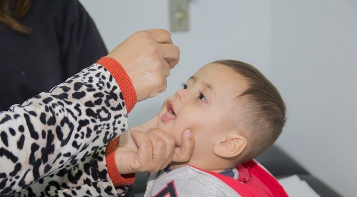 garotinho abre a boca para receber gotas de vacinação, segurado por uma mulher adulta; só se vê os braços e mãos dela