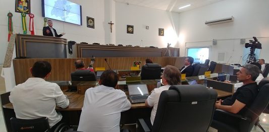 homem fala na tribuna da câmara de palhoça para alguns vereadores sentados, que observam
