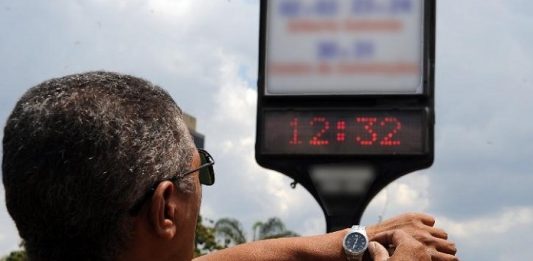 Horário de Verão começará em 4 de novembro homem ajusta relógio de pulso olhando para relógio em painel na rua